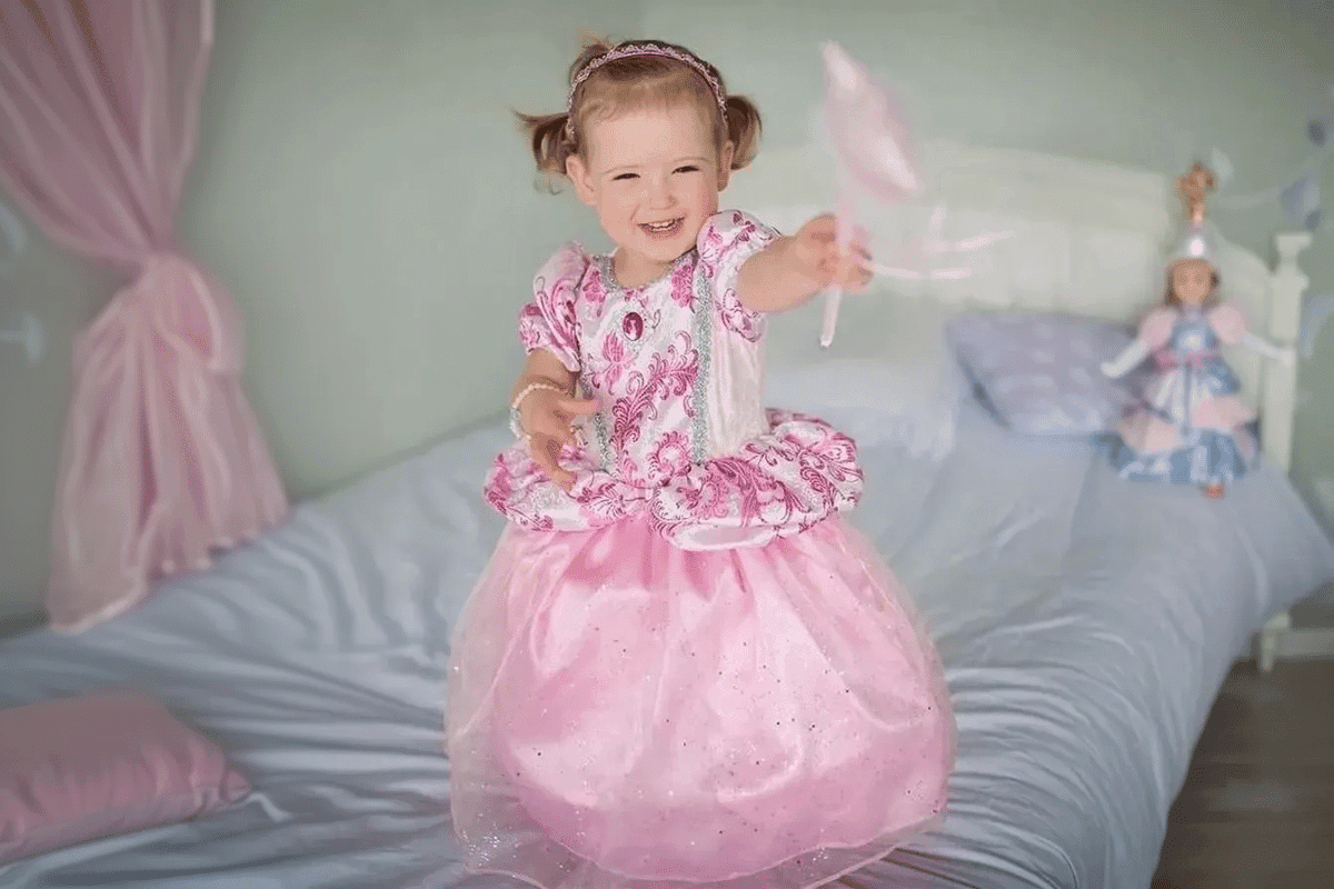 Prinsessekjole. En lille pige står i hendes seng, iført en lyserød prinsessekjole med en tryllestav i hånden.