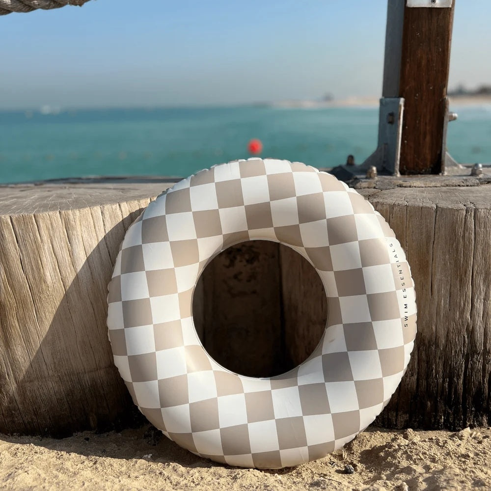 Swim Essential Badering Beige Check liggende ved stranden med havet i baggrunden.